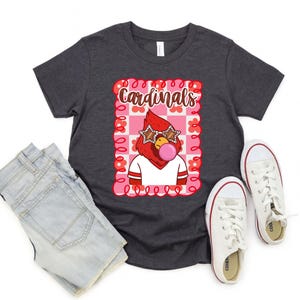 Dark grey heather t-shirt with a graphic of a red cardinal wearing star-shaped sunglasses and blowing a bubblegum bubble. The word "Cardinals" is above the cardinal. White sneakers and denim shorts are next to the shirt.