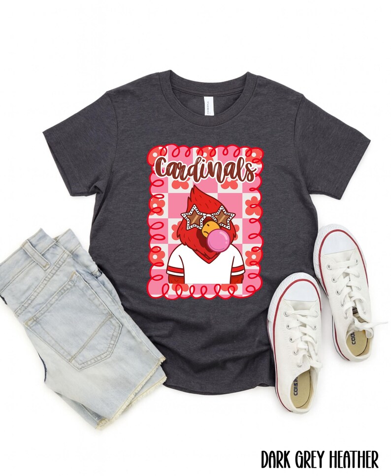 Dark grey heather t-shirt with a graphic of a red cardinal wearing star-shaped sunglasses and blowing a bubblegum bubble. The word "Cardinals" is above the cardinal. White sneakers and denim shorts are next to the shirt.