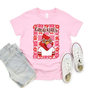 light pink t-shirt featuring a cartoon cardinal with star-shaped sunglasses blowing a bubble. The cardinal is wearing a white shirt with red stripes. The word "Cardinals" is written in brown script within a pink and red floral frame.