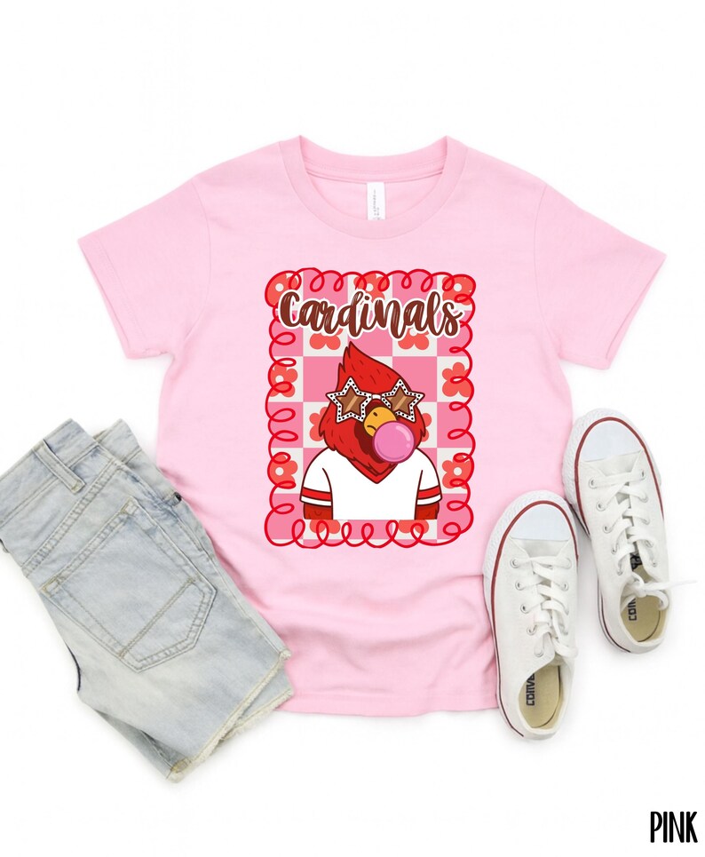 light pink t-shirt featuring a cartoon cardinal with star-shaped sunglasses blowing a bubble. The cardinal is wearing a white shirt with red stripes. The word "Cardinals" is written in brown script within a pink and red floral frame.
