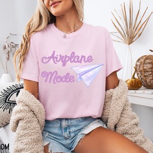 A light pink t-shirt with the words "Airplane Mode" in a purple script font and a paper airplane graphic. The shirt is made of a soft material and is suitable for casual wear.