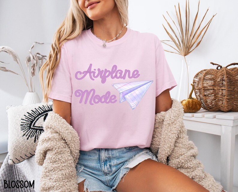 A light pink t-shirt with the words "Airplane Mode" in a purple script font and a paper airplane graphic. The shirt is made of a soft material and is suitable for casual wear.