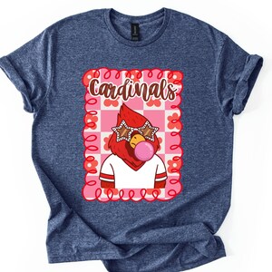 May include: Heather navy blue t-shirt featuring a cartoon cardinal with star-shaped sunglasses blowing a bubblegum bubble. The cardinal is inside a pink and white checkered frame with the word "Cardinals" above it.