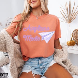Terracotta-colored t-shirt with the words "Airplane Mode" and a paper airplane graphic in purple. The shirt is paired with denim shorts and a beige cardigan. The image is styled with fall-themed decor.