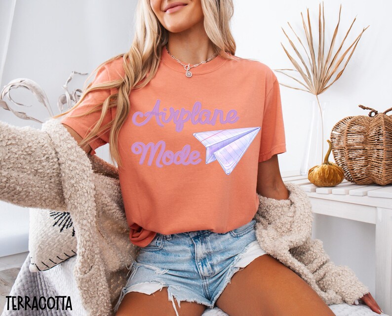 Terracotta-colored t-shirt with the words "Airplane Mode" and a paper airplane graphic in purple. The shirt is paired with denim shorts and a beige cardigan. The image is styled with fall-themed decor.