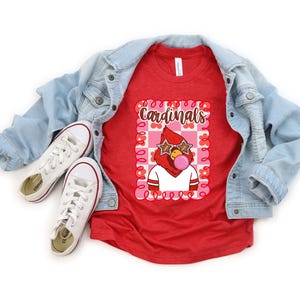 Red t-shirt featuring a cartoon cardinal with star-shaped sunglasses blowing a bubble. The cardinal is wearing a white shirt with red stripes. The word "Cardinals" is written in brown script within a pink and red floral frame.