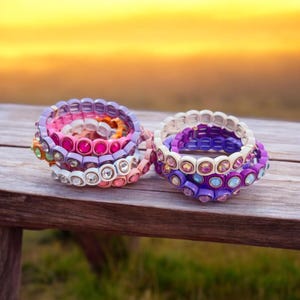 May include: A stack of colorful bracelets with various shades of purple, pink, and white. The bracelets are made of small square beads with rhinestones in the center. The bracelets are arranged on a wooden surface.