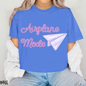 A blue t-shirt with the words "Airplane Mode" in pink script and a paper airplane graphic. The shirt is a solid blue color and has short sleeves.