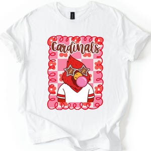 White t-shirt featuring a cartoon cardinal with star-shaped sunglasses blowing a bubble. The cardinal is wearing a white shirt with red stripes. The word "Cardinals" is written in brown script within a pink and red floral frame.
