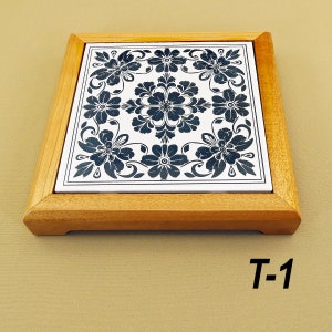 May include: A wooden trivet with a white ceramic tile featuring a black floral design. The tile has a border of black floral designs. The trivet is labeled "T-1".