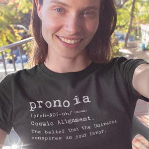 May include: A black short-sleeved t-shirt featuring white text that reads: pronoia /proh-NOY-uh/ (noun) Cosmic Alignment. The belief that the Universe conspires in your favor. The shirt has a crew neckline and a standard fit with a graphic print.