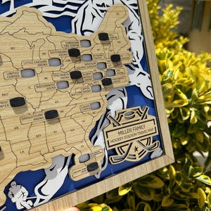 May include: A wooden map of the United States featuring cutouts for hockey arenas, marked with small black puck-shaped tokens. The map includes text labels for various arenas and a plaque reading MILLER FAMILY HOCKEY STADIUM TRAVEL MAP.