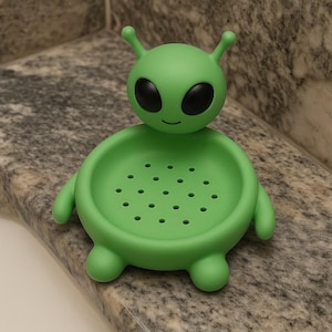 May include: A green alien-shaped soap dish with a smiling face, large black eyes, and small antennae. The dish has a round, perforated base and small arms and feet. The soap dish is made of plastic and is designed to hold a bar of soap.