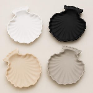Può includere: Quattro piatti a forma di conchiglia in bianco, nero, beige e grigio. Ogni piatto presenta un motivo strutturato a ventaglio che si irradia dal centro. I piatti sono disposti su una superficie beige chiaro, mostrando i loro vari colori e design.