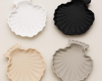 Puede incluir: Cuatro platos de cerámica con forma de concha en blanco, negro, beige y gris. Cada plato tiene una superficie texturizada y acanalada, y un borde festoneado, que se asemeja a una concha real. Los platos están dispuestos sobre un fondo neutro.