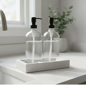 May include: Two clear glass soap or lotion dispensers with black pumps sit on a rectangular concrete tray. The dispensers are filled with a clear liquid. The setup is on a white countertop near a window.