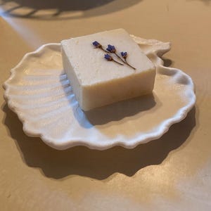 Può includere: Una saponetta quadrata bianco sporco con un rametto di fiori viola è appoggiata su un portasapone bianco a forma di conchiglia. Il portasapone ha un bordo smerlato e una superficie strutturata.