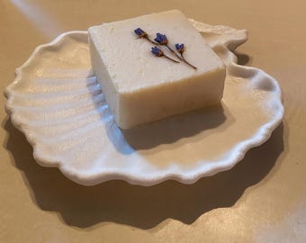 Puede incluir: Una pastilla de jabón cuadrada, blanquecina, con una ramita de flores moradas, descansa sobre una jabonera blanca con forma de concha. La jabonera tiene un borde festoneado y una superficie texturizada.
