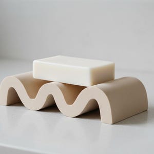 May include: A beige soap dish with a wave-like design, holding a rectangular bar of soap. The soap dish is made of a solid material. The soap is a light cream colour and sits on top of the dish.