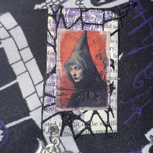May include: A black and white image of a woman wearing a hooded cloak and a pentagram charm with a crescent moon on each side. The image is printed on a piece of paper with a black lace overlay and a pin attached to the top.
