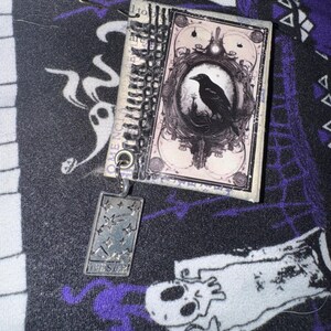 May include: A small, black and white, vintage-style book with a black crow illustration on the cover. The book is attached to a silver charm with the word "The Star" on it. The book and charm are on a black and white fabric background.