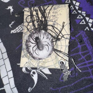 May include: A handmade Halloween charm featuring a black spider on a white background with a black and white spider web design. The charm is attached to a black string and has a silver bat charm hanging from it.