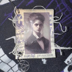 May include: A small, vintage-style card with a black and white portrait of a woman with dark hair and a serious expression. The card has the text "mutual protection" printed on the bottom. A silver pentagram charm is attached to the card with a metal ring.