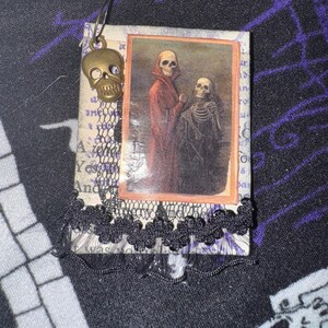 May include: A small, handmade book charm featuring a vintage image of two skeletons. The charm is decorated with black lace and a brass skull pendant.