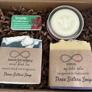 May include: A gift box with handmade soaps and a solid lotion bar. The soaps are labelled "maskihkiwâpoy sweet black tea" and "nimis my older sister bergamot & chamomile." A small tin of "Spearmint" essential oil is also included. The lotion bar has a bee design.