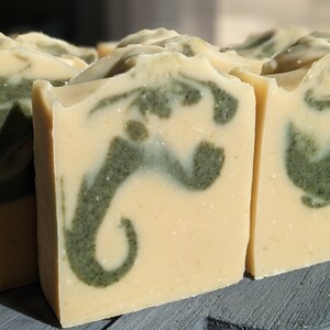 Pode incluir: Close-up de v&aacute;rias barras de sab&atilde;o artesanais. As barras t&ecirc;m uma base amarela cremosa com um padr&atilde;o verde em espiral. O sab&atilde;o &eacute; cortado em formas retangulares e a textura parece lisa e natural.