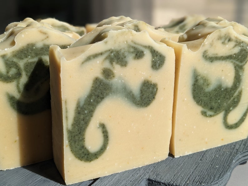 Pode incluir: Close-up de v&aacute;rias barras de sab&atilde;o artesanais. As barras t&ecirc;m uma base amarela cremosa com um padr&atilde;o verde em espiral. O sab&atilde;o &eacute; cortado em formas retangulares e a textura parece lisa e natural.