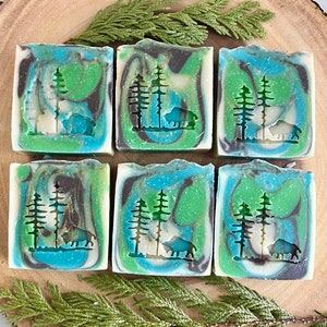 May include: Six bars of handmade soap with a green, blue and white marbled design. Each bar features a silhouette of a pine tree and a bear.
