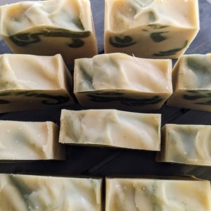 Pode incluir: Uma cole&ccedil;&atilde;o de barras de sab&atilde;o artesanais com uma base cremosa esbranqui&ccedil;ada e redemoinhos verdes. As barras retangulares est&atilde;o dispostas sobre uma superf&iacute;cie escura, mostrando sua apar&ecirc;ncia natural e org&acirc;nica. O sab&atilde;o parece n&atilde;o ter perfume.