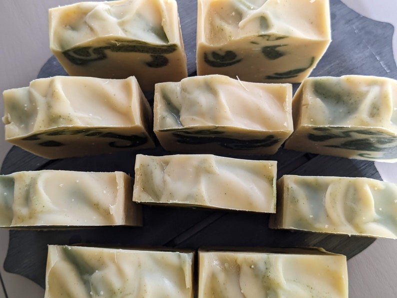 Pode incluir: Uma cole&ccedil;&atilde;o de barras de sab&atilde;o artesanais com uma base cremosa esbranqui&ccedil;ada e redemoinhos verdes. As barras retangulares est&atilde;o dispostas sobre uma superf&iacute;cie escura, mostrando sua apar&ecirc;ncia natural e org&acirc;nica. O sab&atilde;o parece n&atilde;o ter perfume.