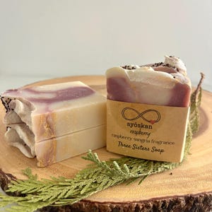 May include: Two bars of handmade soap with a raspberry sangria fragrance, displayed on a wooden slice with greenery. The soap features a layered design with white, pink, and purple tones. The label reads "ayöskan raspberry Three Sisters Soap."
