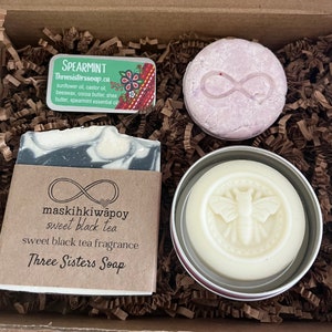 May include: A gift box containing three bath and body products: a bar of black and white marbled soap with the text "maskihkiwapoy sweet black tea sweet black tea fragrance Three Sisters Soap", a round white soap with a bee design, and a round purple shampoo bar with an infinity symbol.