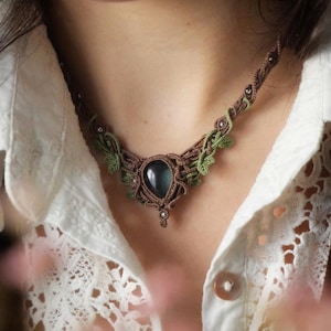 Op de afbeelding: Een handgemaakte macramé ketting met een grote, donkerblauwe edelsteen hanger. De ketting bevat bruin en groen macramé met bladachtige details. De ketting wordt gedragen over een wit kanten shirt.