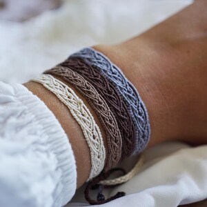 Peut inclure: Trois bracelets tressés dans des tons de gris, marron et blanc, portés au poignet.