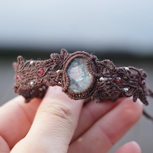 Peut inclure: Un bracelet en macramé marron avec une pierre précieuse verte et blanche et de petites perles rouges.