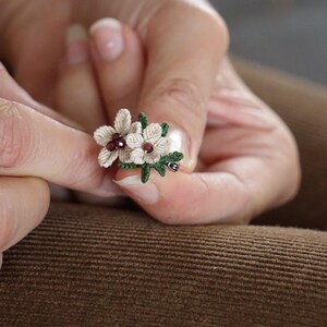 Könnte beinhalten: Eine kleine, zarte Blumenbrosche mit zwei weißen Blüten und grünen Blättern. Die Blüten haben eine burgunderrote Mitte.