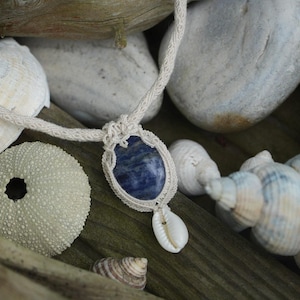 Peut inclure: Un collier artisanal avec un pendentif en pierre bleue dans un cadre macramé, orné d'un petit coquillage. Le collier est présenté avec des coquillages, des roches et du bois flotté, créant un thème côtier naturel.