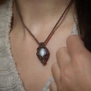 Peut inclure: Un collier artisanal en macramé avec un pendentif central rond et irisé en pierre précieuse. Le pendentif est serti dans un cadre en macramé marron, suspendu à un cordon tressé assorti. Le collier est porté autour du cou.