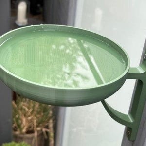 May include: A sage green circular bird bath basin with a smooth interior surface filled with water, attached to a vertical wooden post by a matching green L-shaped mounting bracket secured with two visible metal screws at the top.