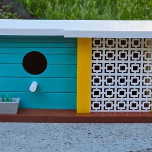 May include: A modern birdhouse with a white roof, turquoise walls, and a yellow accent. The birdhouse features a round entry hole, a small planter box, and a decorative white lattice panel. The base is brown.