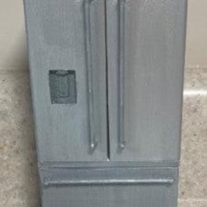 May include: A miniature silver refrigerator with a double-door top and a bottom drawer. The refrigerator has long, vertical handles and a small square dispenser on the left door. The finish has a textured appearance.