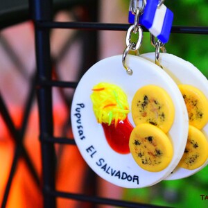 K&ouml;nnte beinhalten: Wei&szlig;er Keramik-Schl&uuml;sselanh&auml;nger mit einem gelben und schwarzen Pupusa-Design. Der Schl&uuml;sselanh&auml;nger tr&auml;gt den Text "Pupusas El Salvador".