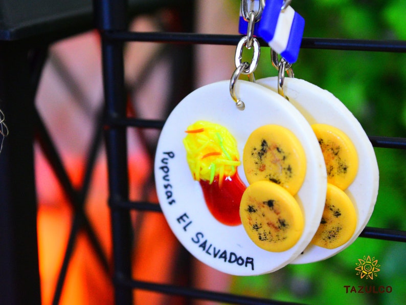 K&ouml;nnte beinhalten: Wei&szlig;er Keramik-Schl&uuml;sselanh&auml;nger mit einem gelben und schwarzen Pupusa-Design. Der Schl&uuml;sselanh&auml;nger tr&auml;gt den Text "Pupusas El Salvador".