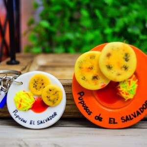 Puede incluir: Dos llaveros con platos en miniatura de pupusas, un plato tradicional salvadore&ntilde;o. Los llaveros son blancos y naranjas con el texto "Pupusas de El Salvador" en cada uno.