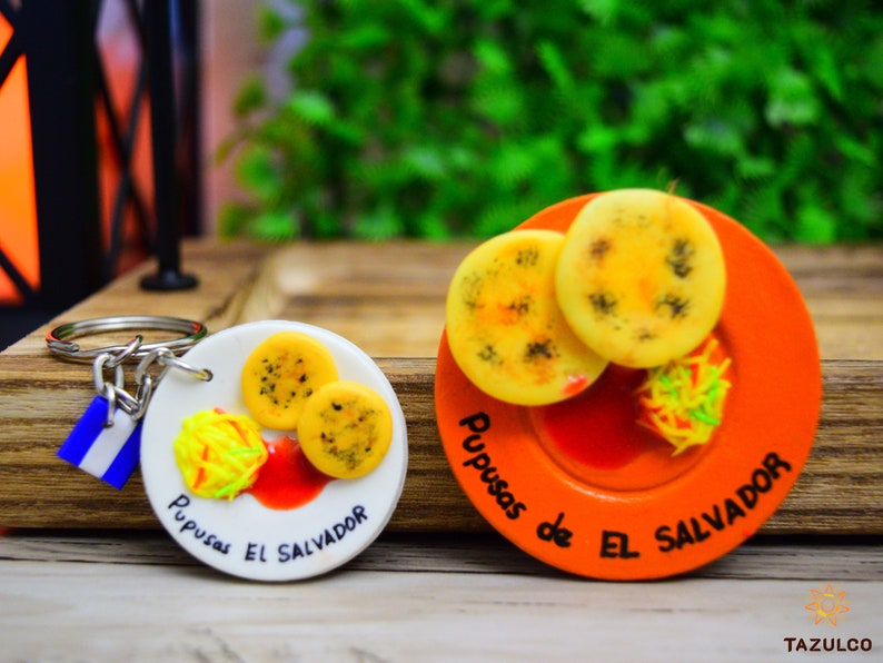 Puede incluir: Dos llaveros con platos en miniatura de pupusas, un plato tradicional salvadore&ntilde;o. Los llaveros son blancos y naranjas con el texto "Pupusas de El Salvador" en cada uno.