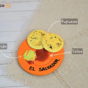 Puede incluir: Primer plano de un peque&ntilde;o plato naranja con las palabras "El Salvador" escritas en &eacute;l. El plato tiene dos pupusas amarillas, un poco de salsa roja y un poco de curtido verde. El plato est&aacute; sobre un fondo de arpillera beige.